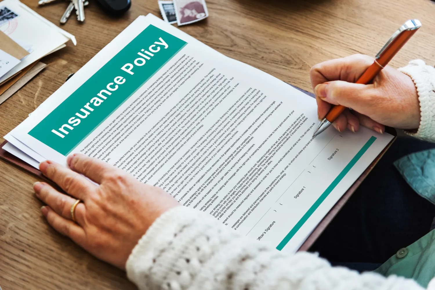 A person signing an insurance policy document with a pen on a wooden table, with additional papers and keys nearby.